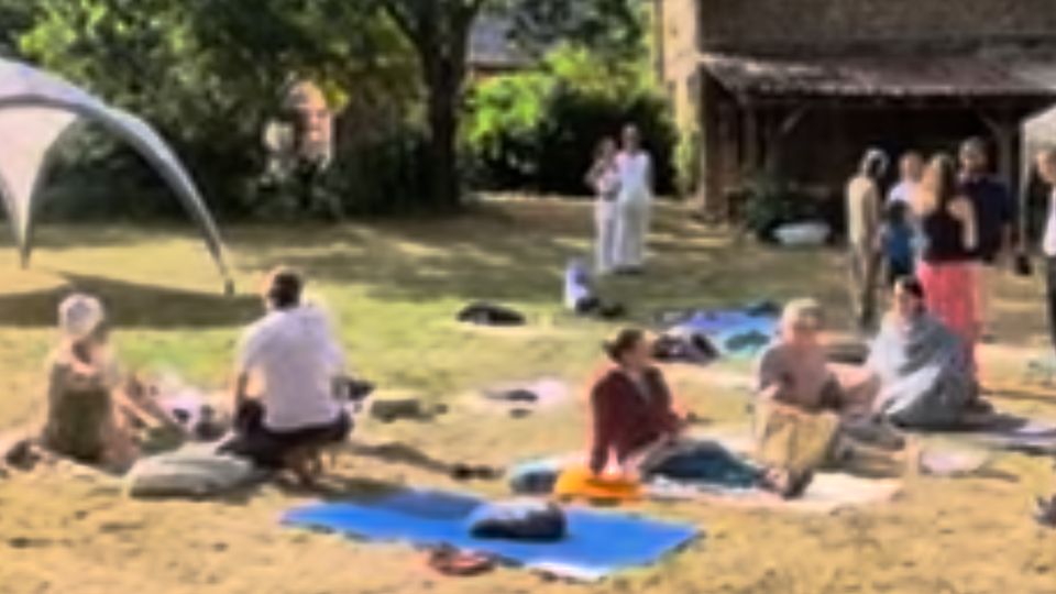 Menschen sitzen und stehen auf einer Wiese vor Haus Bollheim, auf Decken und Matten, unter Bäumen in der Sonne.