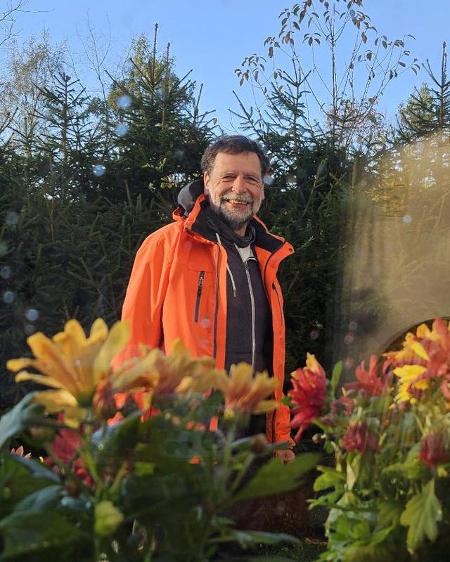 Bernd Marnau lächelt in einer orangefarbenen Jacke, vor Tannen und hinter Herbstblumen im Garten.