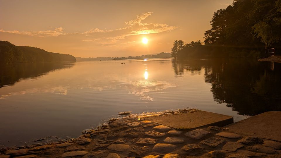 Sonnenuntergang über dem ruhigen See in Bad Segeberg, die Sonne spiegelt sich im Wasser, rechts und links bewaldetes Ufer.